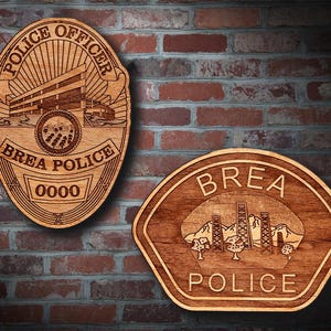 May include: Two wooden Brea Police badges. One is oval-shaped with "POLICE OFFICER" at the top and "BREA POLICE" at the bottom. The other is a shield shape with "BREA POLICE" at the top and "POLICE" at the bottom. Both are brown.