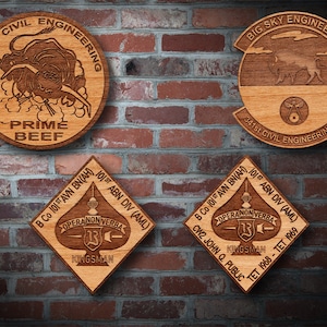 May include: Four wooden plaques with engraved designs. The top left plaque is round and features a bull with the text "USAF CIVIL ENGINEERING PRIME BEEF". The top right plaque is round and features a buffalo with the text "BIG SKY ENGINEERS 341st CIVIL ENGINEERING SO". The bottom left plaque is diamond shaped and features a spade with the text "B Co 101 ST AVN BN(AH) 101ST ABN DIV (AML) OPERA NON VERBA KINGSMAN". The bottom right plaque is diamond shaped and features a spade with the text "B Co 101 ST AVN BN(AH) 101ST ABN DIV (AML) OPERA NON VERBA KINGSMAN CW2 JOHN Q. PUBLIC TET 1968-TET 1969".