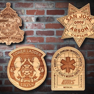 May include: Four wooden plaques with engraved designs. The top left plaque features a crest with the text "Kanaka Fire & Emergency Services". The top right plaque is a six-pointed star with the text "San Jose 000F Arson Captain". The bottom left plaque features two lions with the text "Tower Ladder FD XL119 NY Lions". The bottom right plaque features a star of life with the text "Robert Wood Johnson University Hospital St. Peter's Medical Center Emergency EMS Medical Service".