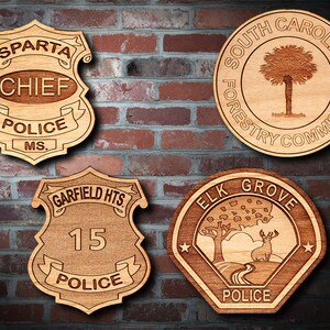 May include: Four wooden plaques with engraved designs. The top left plaque is a police badge with the words "SPARTA CHIEF POLICE MS.". The top right plaque is a round plaque with the words "SOUTH CAROLINA FORESTRY COMMISSION" and a palm tree design. The bottom left plaque is a police badge with the words "GARFIELD HTS. 15 POLICE". The bottom right plaque is a hexagonal plaque with the words "ELK GROVE POLICE" and a deer in a forest scene.