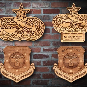 May include: Wooden plaques with military emblems. Two plaques feature an eagle holding a weapon, with a star at the top. The other two plaques have a shield design with the text "AIR FORCE MEDICAL READINESS AGENCY" and the name "Sgt. John Q. Public".