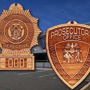 May include: Two wooden law enforcement badges. One is a starburst shape with the text "Passaic County Prosecutor's Office, State of New Jersey, Detective, 000". The other is a shield shape with "Prosecutor's Office, Passaic County, New Jersey, 1837".