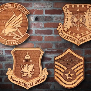 May include: Four wooden plaques with laser-engraved designs. The top left plaque features a flying eagle with the text "Sweat More, Bleed Less" and "966 AACS". The top right plaque features a shield with crossed arrows and the text "USAF Expeditionary Center". The bottom left plaque features a lion with a building in the background and the text "316th Medical Group". The bottom right plaque features a chevron with a star and the text "MSGT John Q. Public 0000".