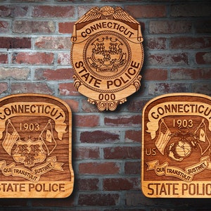 May include: Three wooden plaques featuring the Connecticut State Police logo. The top plaque displays the words "Connecticut State Police" and "000" within a circle. The left plaque shows the words "Connecticut State Police" and "1903" alongside the state seal and flags. The right plaque features the words "Connecticut State Police" and "1903" with the US and Marine Corps flags.