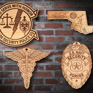 Könnte beinhalten: Vier Holzplaketten mit lasergravierten Designs. Die obere linke Plakette zeigt eine Justizwage, einen Blitz und ein Wappen mit einer Lilie. Der Text lautet "To Serve With Honor 7100 Security Police SQ". Die obere rechte Plakette ist eine Karte von Maryland mit dem Text "GMI Joseph White Senior Masterchief, National Capital Region June 2017 - May 2019". Die untere linke Plakette zeigt einen Caduceus mit Flügeln. Die untere rechte Plakette zeigt ein Abzeichen mit dem Text "U.S. Navy Security Forces" und einem Stern.