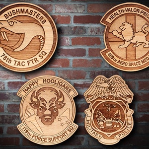 May include: Four wooden plaques with engraved military unit insignia. The top left plaque features a snake with the text "BUSHMASTERS 78th TAC FTR SQ". The top right plaque features a lion with the text "HEALTH-VALOR-PRIDE 459th AERO SPACE MED SQ". The bottom left plaque features a bull with the text "HAPPY HOOLIGANS 119th FORCE SUPPORT SQ". The bottom right plaque features an eagle with the text "UNITED STATES ARMY MILITARY POLICE".
