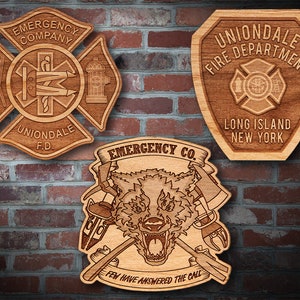 Könnte beinhalten: Drei Holzplaketten mit Feuerwehr-Motiven. Die obere Plakette ist schildförmig mit dem Text "Uniondale Fire Department Long Island New York". Die mittlere Plakette ist ein Kreuz mit dem Text "Emergency Company Uniondale F.D.". Die untere Plakette zeigt einen Wolf mit gekreuzten Äxten und dem Text "Emergency Co. Few Have Answered The Call."