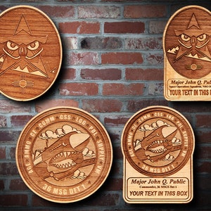 May include: Four wooden plaques with laser-engraved designs. The top left plaque features an owl with a mountain range and a sun symbol. The top right plaque features an owl with a mountain range and the text "Major John Q. Public Space Operations Squadron, NRO Delta 23 YOUR TEXT IN THIS BOX". The bottom left plaque features a plane with the text "AGE AMMO CE COMM+CSS+LOG POL SUPPLY W/N 36 MSG DET 1". The bottom right plaque features a plane with the text "AGE AMMO CE COMM+CSS+LOG POL SUPPLY W/N 36 MSG DET 1 Major John Q. Public Commander, 36 MSGS Det 1 YOUR TEXT IN THIS BOX".