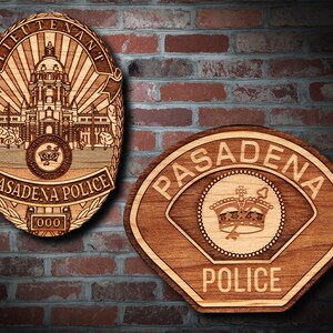 Könnte beinhalten: Zwei hölzerne Polizeibadgen, einer mit den Worten "PASADENA POLICE" und einer Krone, der andere mit den Worten "LIEUTENANT" und "PASADENA POLICE" und einem detaillierten Bild eines Gebäudes.