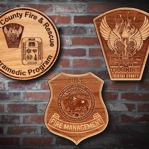 May include: Three wooden plaques with engraved designs. The top left plaque is round and features the text "Fairfax County Fire & Rescue Paramedic Program". The top right plaque is shaped like a shield and features the text "EMS Training Fairfax County". The bottom plaque is shaped like a shield and features the text "U.S. Department of the Interior Bureau of Land Management Fire Management".