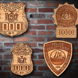Könnte beinhalten: Vier hölzerne Polizeibadges mit unterschiedlichen Designs. Das obere linke Abzeichen zeigt "Port Chester Police" mit einem Siegel des Staates New York und der Nummer "000". Das obere rechte Abzeichen zeigt "Port Chester Police Detective" mit einem Siegel des Staates New York und der Nummer "0000". Das untere linke Abzeichen zeigt "Sergeant Port Chester Police" mit einem Siegel des Staates New York und der Nummer "000". Das untere rechte Abzeichen zeigt "Police Department Port Chester 1868 New York" mit einem Schiff und dem Text "Police Department".