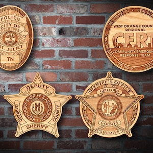 May include: Four wooden plaques with engraved designs of law enforcement badges. The top left plaque reads "Police City of Mt. Juliet TN". The top right plaque reads "West Orange County Regional CERT Community Emergency Response Team". The bottom left plaque reads "Deputy Garrard Kentucky Sheriff's Association County Sheriff". The bottom right plaque reads "Sheriff's Office Guilford County Sheriff".