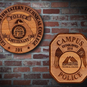 May include: Two wooden signs with the text "Georgia Northwestern Technical College Police" and "Campus Established 2018". The signs are both brown and have a wood grain texture. The first sign is round and has a star on each side of the number "101". The second sign is octagonal and has a stylized image of two American flags crossed over a mountain range.