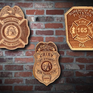 May include: Three wooden wall plaques shaped like fire department badges. The top left badge reads Station Chief State of Georgia GDC Fire Services. The top right badge reads Bureau of Fire Pittston City Fire 165 Rescue PA. The bottom badge reads Retired Chief Dept of Forestry Fire Protection 0000.