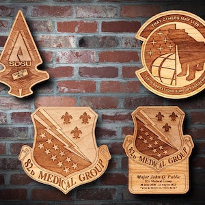 May include: Four wooden plaques with laser-engraved military unit insignia. The top plaque features a bear and the text "THAT OTHERS MAY LIVE 129TH OPERATIONS SUPPORT SQUADRON". The bottom left plaque features a lightning bolt and fleur-de-lis with the text "82D MEDICAL GROUP". The bottom right plaque features a lightning bolt and fleur-de-lis with the text "82D MEDICAL GROUP" and a personalized message below. The top right plaque features a stylized arrow with the text "TIP OF THE SPEAR COMMANDANT'S AWARD SDSU YOUR TEXT HERE".