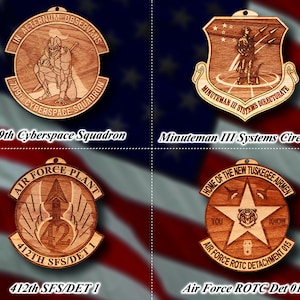 May include: Four wooden ornaments with different designs. The top left ornament features a knight in armor with the text "IN AETERNUM OBSERVANS 709TH CYBERSPACE SOUADRON". The top right ornament features a soldier with the text "MINUTEMAN III SYSTEMS DIRECTORATB". The bottom left ornament features a rocket with the text "AIR FORCE PLANT 42 412TH SFS/DET 1". The bottom right ornament features a star with a tiger inside and the text "HOME OF THE NEW TUSKEGEE AIRMEN YOU KNOW AIR FORCE ROTC DETACHMENT 015".