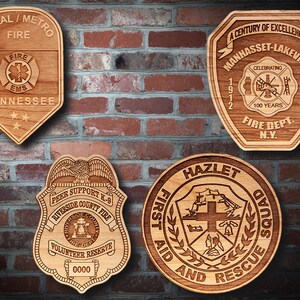 May include: Four wooden plaques with engraved designs. The top left plaque is a brown, teardrop-shaped plaque with the text "Rural/Metro Fire Tennessee" and a fire department logo. The top right plaque is a brown, octagonal plaque with the text "A Century of Excellence Manhasset-Lakeville Fire Dept. N.Y." and a fire department logo. The bottom left plaque is a brown, shield-shaped plaque with the text "Peer Support K-9 Riverside County Fire Volunteer Reserve 0000" and a fire department logo. The bottom right plaque is a brown, round plaque with the text "Hazlet First Aid and Rescue Squad" and a fire department logo.
