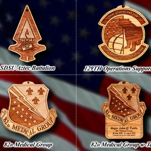 May include: Four wooden ornaments with different designs. The first ornament is a brown spear with the text "SDSU" and "YOUR TEXT HERE". The second ornament is a brown circle with a bear and the text "THAT OTHERS MAY LIVE" and "129TH OPERATIONS SUPPORT SQUADRON". The third ornament is a brown shield with a lightning bolt and the text "82D MEDICAL GROUP". The fourth ornament is a brown shield with a lightning bolt and the text "82D MEDICAL GROUP" and "Major John Q. Public 82D Medical Group 20 June 2020 - 23 August 2023 "YOUR TEXT IN THIS BOX"