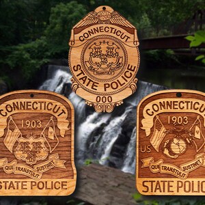 May include: Three wooden ornaments featuring the Connecticut State Police logo. The top ornament is a traditional shield design with the words "Connecticut State Police" and "000". The two bottom ornaments are rectangular with the words "Connecticut State Police" and "1903".