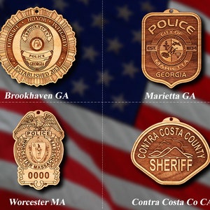 May include: Four wooden ornaments featuring police and sheriff badges. The top left ornament is a round badge with the words "Courage, Honor, Integrity" and "Brookhaven Police Georgia Established 2013". The top right ornament is a shield-shaped badge with the words "Police City of Marietta Georgia Est. 1834". The bottom left ornament is a badge with the words "Police Worcester Massachusetts" and a number "0000". The bottom right ornament is a shield-shaped badge with the words "Contra Costa County Sheriff".
