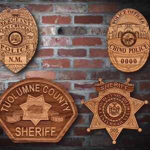 May include: Four wooden police and sheriff badges with engraved details. The badges are in the shape of a star, a shield, and a circle. The badges have text that reads "Las Cruces Police", "Chino Police", "Tuolumne County Sheriff", and "Ulster County Sheriff".