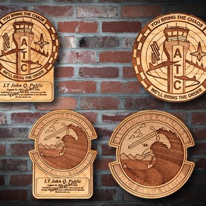 May include: Four wooden plaques with engraved designs. Two plaques feature a circular design with the text "You bring the chaos, A T C, We'll bring the order". The other two plaques feature a design of a bird with a sword and the text "AETC Studies & Analysis SQ, Vincit Omnia Veritas". Each plaque has the text "LT John Q. Public" and a date.
