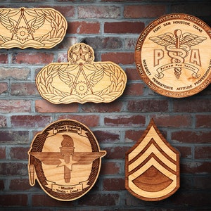 May include: Five wooden plaques with laser-engraved designs. The top left plaque features a military insignia with wings and a gear. The top right plaque features a circle with the text "INTERSERVICE PHYSICIAN ASSISTANT PROGRAM" and "PSA" with a medical symbol. The bottom left plaque features a circle with the text "Military Training Leader School" and a torch with wings. The bottom right plaque features a chevron with three stripes. The center plaque features a star with five points.
