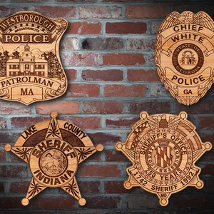 May include: Four wooden police and sheriff badges with engraved designs. The badges are displayed on a brick wall. The top left badge reads "Westborough Massachusetts Police Patrolman MA". The top right badge reads "Chief White State of Georgia Police GA". The bottom left badge reads "Lake County Sheriff Indiana". The bottom right badge reads "Sheriff's Office 250 Years 1742 Sheriff 1992 MD".