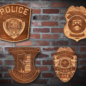 May include: Four wooden plaques with engraved police badges. The top left plaque reads "Police Forks Township Pennsylvania Est. 1754" and features a shield with a mountain and a tree. The top right plaque reads "United States Treasury Controller of the Currency Treasury 2886" and features a shield with a scale of justice and a key. The bottom left plaque reads "Emergency Operations One Team One Mission Security and Emergency Management EOU" and features a shield with a bird, a lighthouse, and a lightning bolt. The bottom right plaque reads "Captain Police 114" and features a shield with an eagle and a star.
