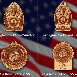 May include: Four wooden ornaments with different designs. The top left ornament is a police badge with the words "Police Officer" and "Arlington County". The top right ornament is a police badge with the words "Police Officer" and "0000". The bottom left ornament is a circular ornament with the words "Tennessee Valley Authority Browns Ferry Nuclear Security". The bottom right ornament is a rectangular ornament with the words "Tennessee Valley Authority Browns Ferry Nuclear Security" and "2023 Security Team of the Year A-Team".
