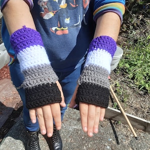 May include: A pair of crocheted wrist warmers in the colors of the asexual pride flag: purple, white, gray, and black.