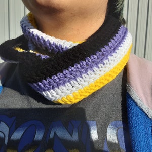 Puede incluir: Un calentador de cuello de ganchillo en rayas amarillas, blancas, moradas y negras. Los colores representan la bandera del orgullo no binario.