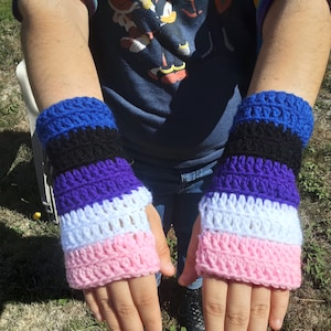 Op de afbeelding: Gehaakte vingerloze handschoenen in een gestreept patroon van blauw, zwart, paars, wit en roze. De handgemaakte polswarmers worden gepresenteerd tegen een natuurlijke buitenachtergrond. De handschoenen zijn een uniek accessoire.