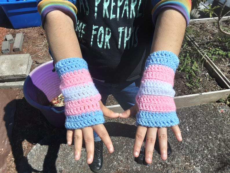 May include: A pair of crocheted wrist warmers in pink, white, and blue. The wrist warmers are striped with the colors of the transgender pride flag.