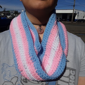 May include: A crocheted infinity scarf with blue, pink, and white stripes. The scarf is worn by a person with a necklace featuring a crystal pendant.