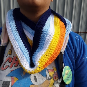 May include: A person wearing a blue jacket and a white t-shirt with a cartoon character on it. They are wearing a crocheted scarf with stripes of blue, white, light blue, yellow, and orange. The scarf is worn around the neck and is folded in half. The person is also wearing a green button with the text "They/Them" on it.