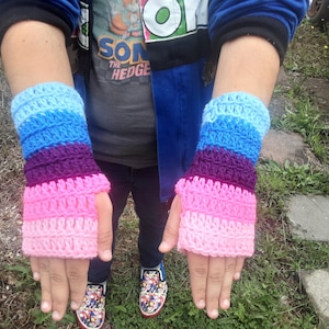 May include: A pair of crocheted fingerless gloves with a pink, purple, blue, and light blue color scheme. The gloves are worn by a person with a blue jacket and a Sonic the Hedgehog t-shirt.