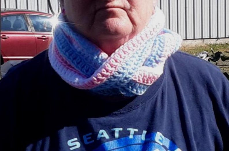 May include: A person wearing a blue shirt with the word "Seattle" on it. They are wearing a crocheted neck warmer with pink, blue, and white stripes.