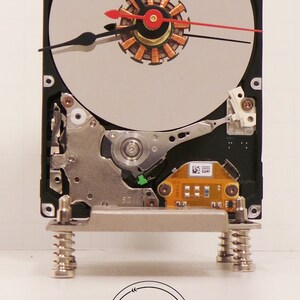May include: A black hard drive clock with a silver face and red and black hands. The clock is mounted on two silver springs. The clock face has a copper motor in the center. The clock is labeled "D2 Distressed Design".