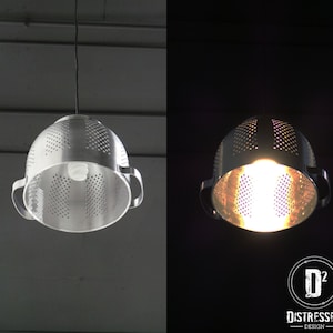 May include: A unique pendant light fixture made from a repurposed metal colander. The silver-colored colander has numerous small holes and two handles. A light bulb is visible inside, casting a warm glow. The fixture hangs from a black cord.