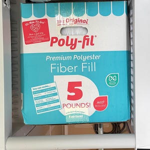 May include: A box of Poly-fil Premium Polyester Fiber Fill weighing 5 pounds. The packaging is light blue and white with red text, featuring a handle cutout at the top, a We Make For Good logo, and a chart detailing project quantities.