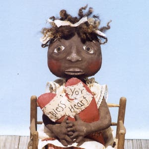 May include: A handmade rag doll with brown hair and a brown face, wearing a white dress with red and white stripes, holding a red heart pillow with the text "Bless Yo'r Heart" embroidered on it. The doll is sitting on a wooden chair.