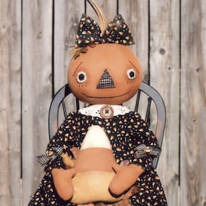 May include: A handmade pumpkin doll wearing a black and white dress with a candy corn design. The doll is holding a candy corn and is sitting on a wooden chair.