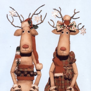 Op de afbeelding: Twee handgemaakte ragdoll rendieren, een met een rood-witte kerstmanmuts en de andere met een rode jurk met een geruite rok en een rode strik. Beide rendieren hebben bruine gewei, bruine ogen en bruine neuzen. Ze zitten op een rood stoffen oppervlak.