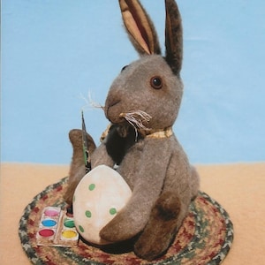 May include: A grey plush bunny rabbit sits on a brown and green braided rug, holding a white egg with green polka dots. A small paint palette with colourful paints is in front of the bunny.