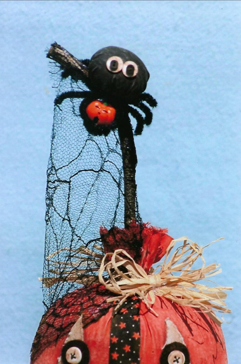 Puede incluir: Una decoraci&oacute;n de calabaza de Halloween hecha a mano con una ara&ntilde;a negra en la parte superior, una tela estampada en blanco y negro y un lazo de paja marr&oacute;n.