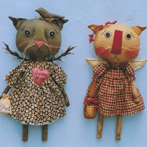 May include: Two handmade cat dolls with painted faces and fabric bodies. One doll is wearing a black and white floral dress with a pink heart that says "Garden Angel" and is holding a small bag that says "Seed". The other doll is wearing a red and black plaid dress and has a white halo. Both dolls have twig legs.