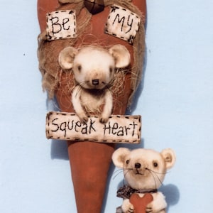 May include: A red heart-shaped fabric with a brown burlap bow and the words "Be My Squeak Heart" on it. Two white felt mice are attached to the heart, one holding a sign and the other holding a red heart.