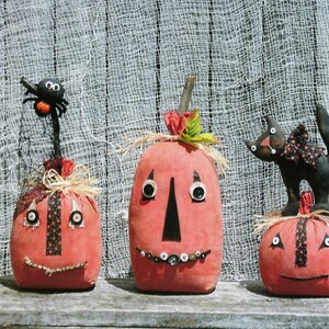 Puede incluir: Tres calabazas de tela hechas a mano con ojos y bocas negros. Las calabazas est&aacute;n decoradas con botones y recortes de tela. Una calabaza tiene una ara&ntilde;a negra en la parte superior.