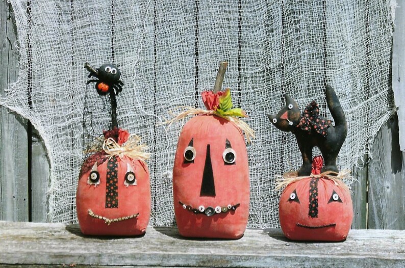 Puede incluir: Tres calabazas de tela hechas a mano con ojos y bocas negros. Las calabazas est&aacute;n decoradas con botones y recortes de tela. Una calabaza tiene una ara&ntilde;a negra en la parte superior.